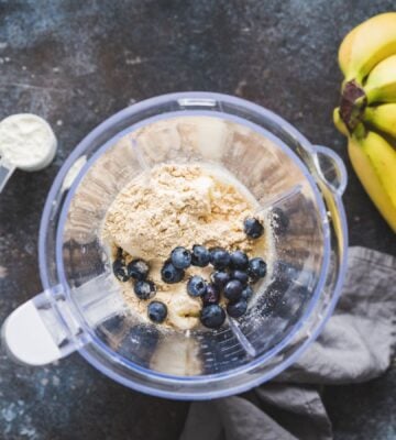 Photo shows an open blender from above containing blueberries and vegan protein powder next to a bunch of a bananas