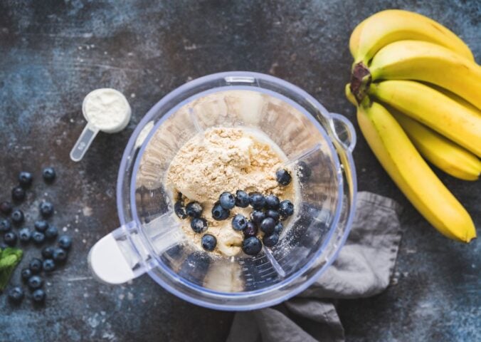 Photo shows an open blender from above containing blueberries and vegan protein powder next to a bunch of a bananas
