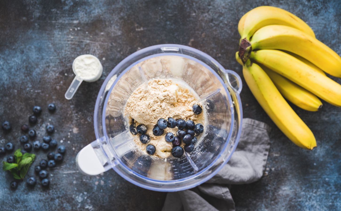 Photo shows an open blender from above containing blueberries and vegan protein powder next to a bunch of a bananas