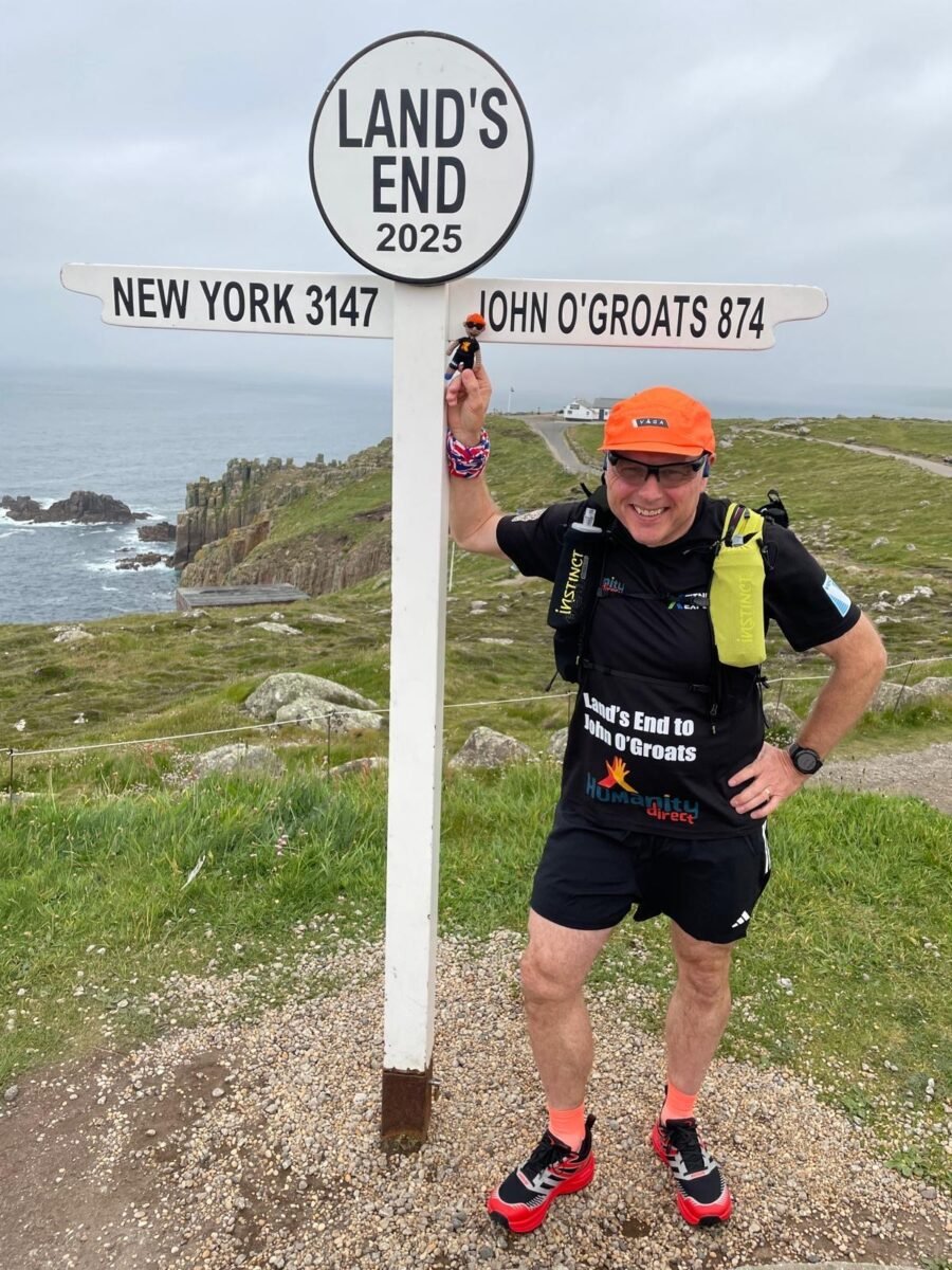 Photo shows vegan runner Bob Yates posing with the sign at Land's End that shows 874 miles to John O'Groats