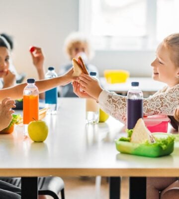 Photo shows a group of school children eating lunch - including juice, fruit, and vegetables - around a cafeteria table
