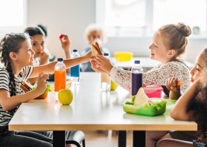 Photo shows a group of school children eating lunch - including juice, fruit, and vegetables - around a cafeteria table