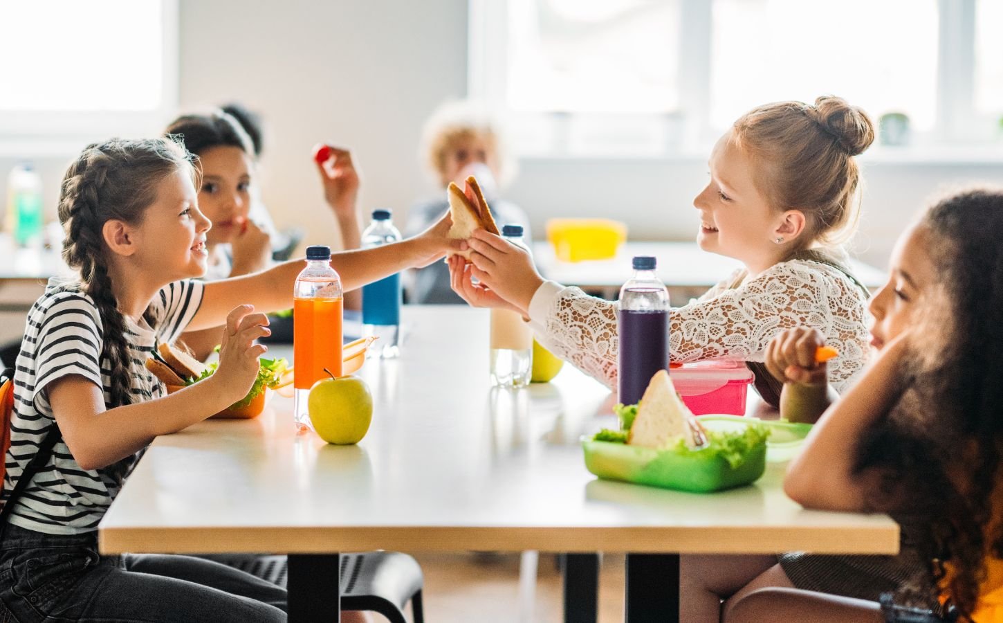 Photo shows a group of school children eating lunch - including juice, fruit, and vegetables - around a cafeteria table
