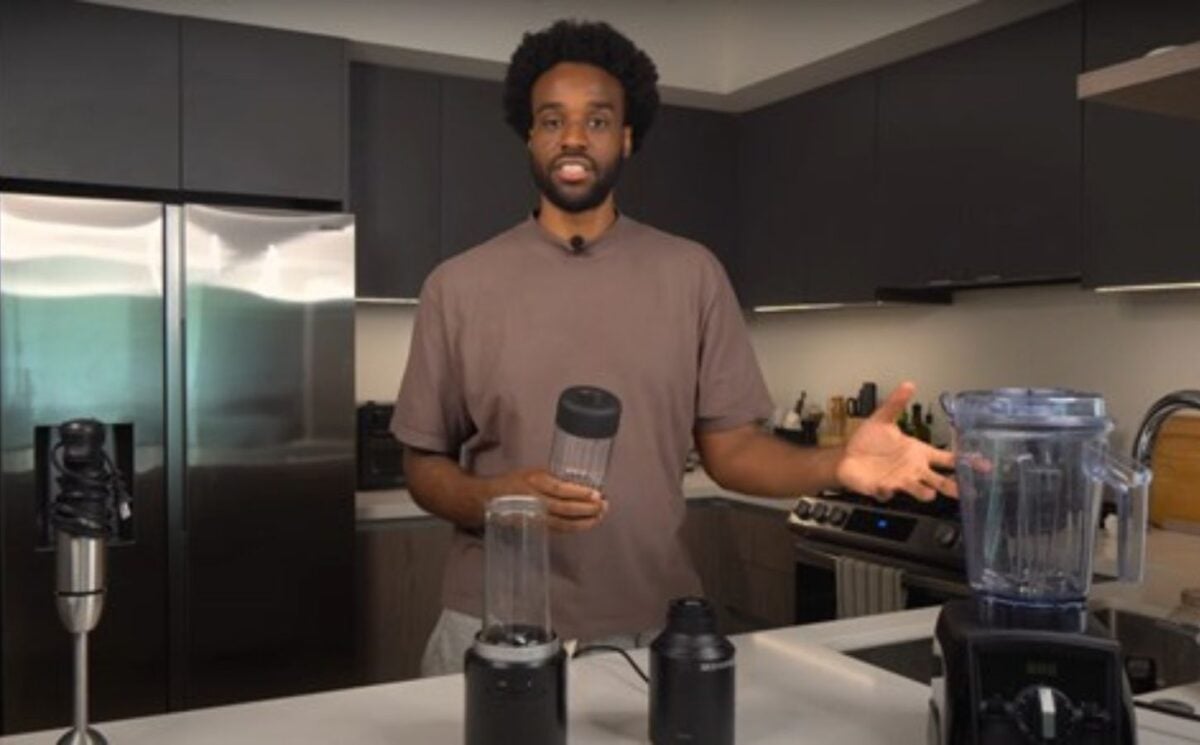 Malcolm Regisford stands in a kitchen holding a personal blender, surrounded by blending appliances on a counter