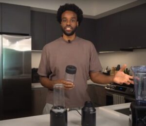 Malcolm Regisford stands in a kitchen holding a personal blender, surrounded by blending appliances on a counter