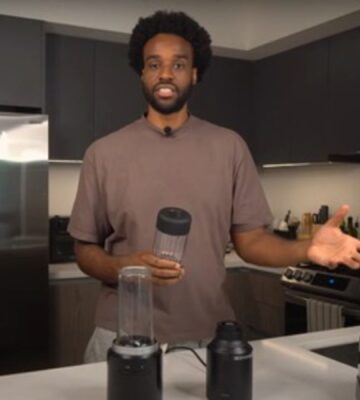 Malcolm Regisford stands in a kitchen holding a personal blender, surrounded by blending appliances on a counter