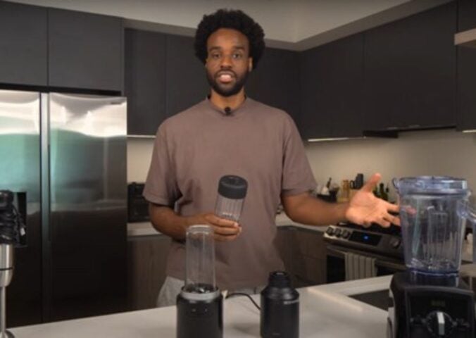 Malcolm Regisford stands in a kitchen holding a personal blender, surrounded by blending appliances on a counter