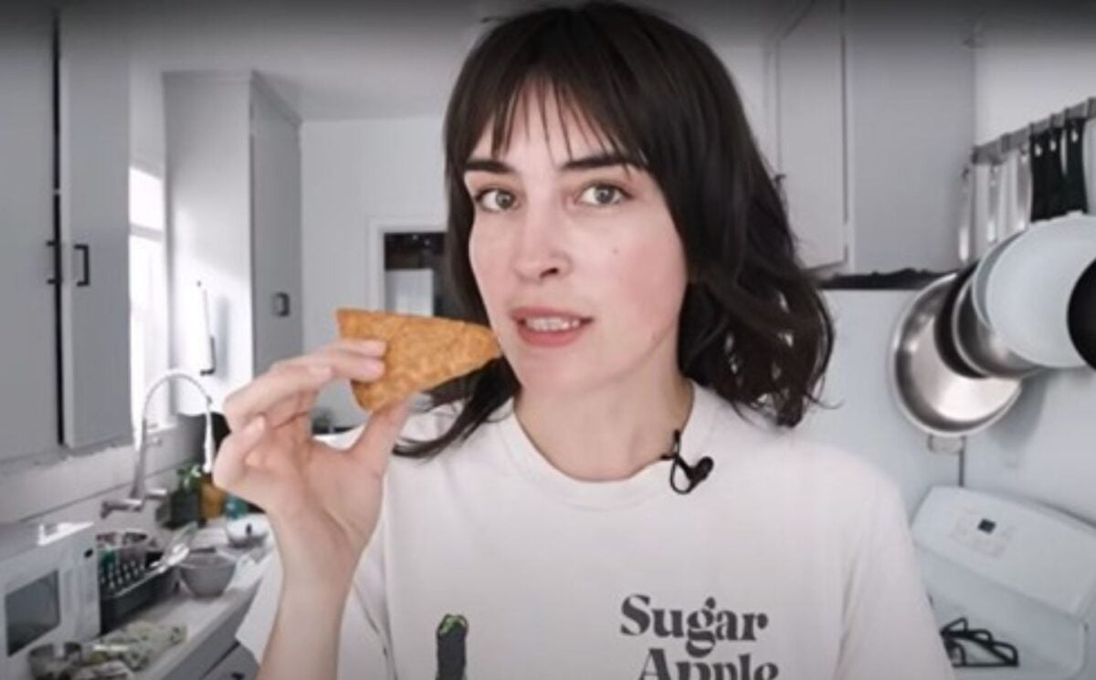 Vegan YouTuber Merle O'Neal holds a wedge of tempeh parmesan that's ready to be grated as she stands in her kitchen