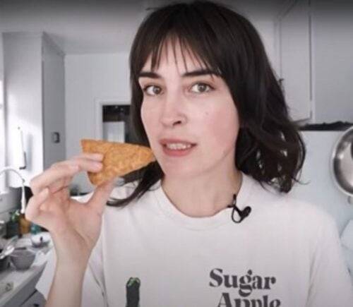 Vegan YouTuber Merle O'Neal holds a wedge of tempeh parmesan that's ready to be grated as she stands in her kitchen