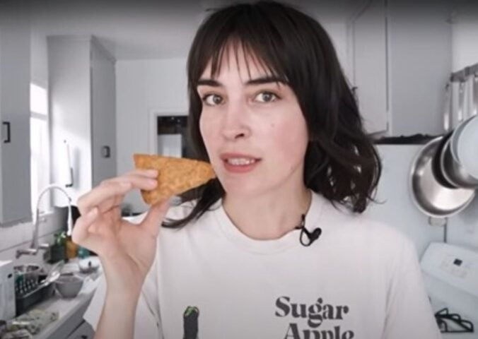 Vegan YouTuber Merle O'Neal holds a wedge of tempeh parmesan that's ready to be grated as she stands in her kitchen