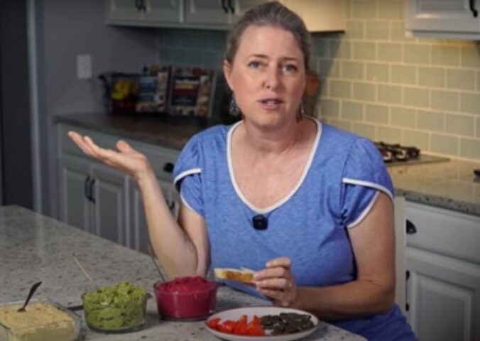 Jill Dalton presents three different oil-free hummus recipes while holding a piece of toast in a kitchen