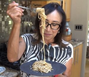 Miyoko Schinner pulls a forkful of her freshly cooked vegan cacio e pepe up from the black plate she's holding in her hand as she stands in her kitchen