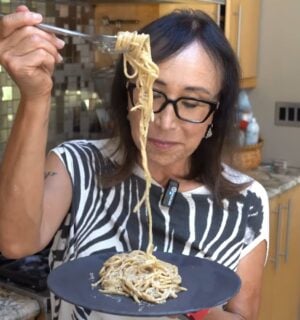 Miyoko Schinner pulls a forkful of her freshly cooked vegan cacio e pepe up from the black plate she's holding in her hand as she stands in her kitchen