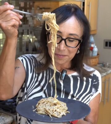 Miyoko Schinner pulls a forkful of her freshly cooked vegan cacio e pepe up from the black plate she's holding in her hand as she stands in her kitchen