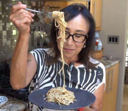 Miyoko Schinner pulls a forkful of her freshly cooked vegan cacio e pepe up from the black plate she's holding in her hand as she stands in her kitchen