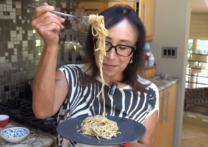 Miyoko Schinner pulls a forkful of her freshly cooked vegan cacio e pepe up from the black plate she's holding in her hand as she stands in her kitchen