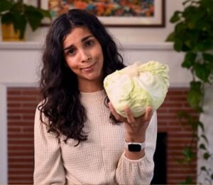 Vegan chef Nisha Vora holds a head of cabbage with a skeptical look on her face as she explores ways to overcome veggie aversion