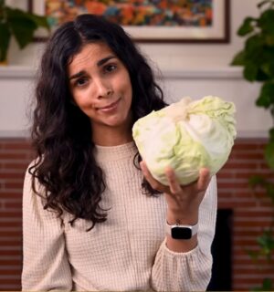 Vegan chef Nisha Vora holds a head of cabbage with a skeptical look on her face as she explores ways to overcome veggie aversion