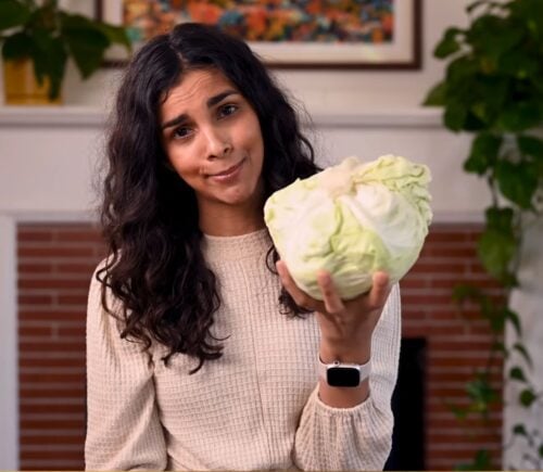 Vegan chef Nisha Vora holds a head of cabbage with a skeptical look on her face as she explores ways to overcome veggie aversion