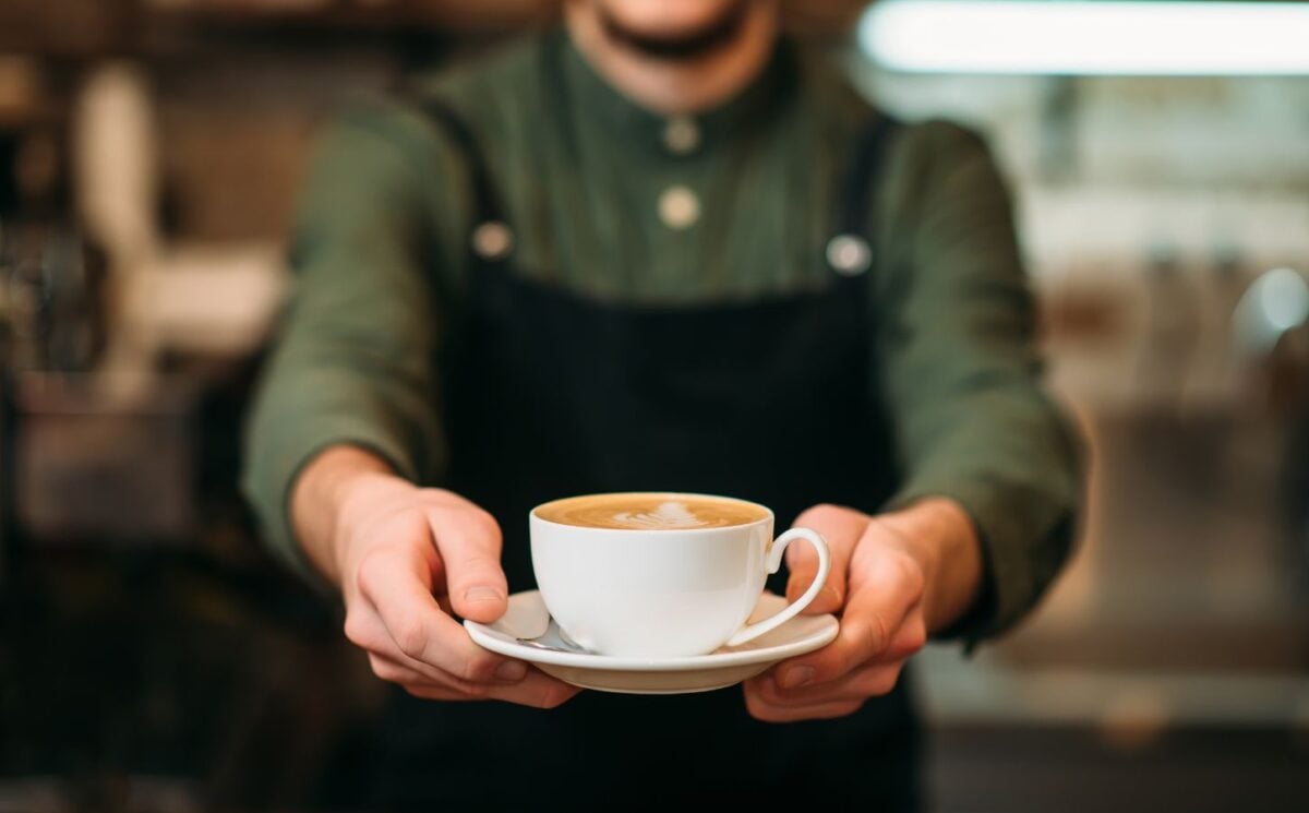 Photo shows a barista holding out a cup of coffee in a saucer