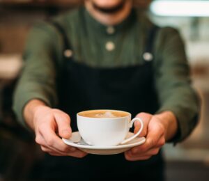 Photo shows a barista holding out a cup of coffee in a saucer