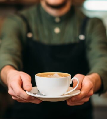 Photo shows a barista holding out a cup of coffee in a saucer