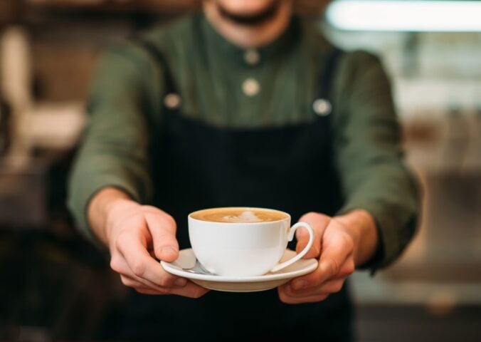 Photo shows a barista holding out a cup of coffee in a saucer