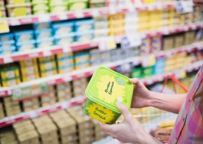 Photo shows a woman holding up a tub of butter-style margarine spread in a supermarket