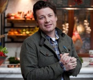 Photo shows Jamie Oliver standing in front of a kitchen stocked with fresh fruit and vegetables