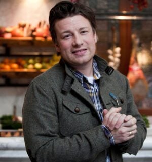 Photo shows Jamie Oliver standing in front of a kitchen stocked with fresh fruit and vegetables