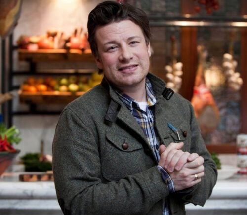 Photo shows Jamie Oliver standing in front of a kitchen stocked with fresh fruit and vegetables