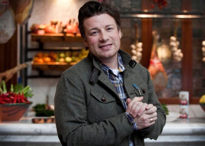 Photo shows Jamie Oliver standing in front of a kitchen stocked with fresh fruit and vegetables