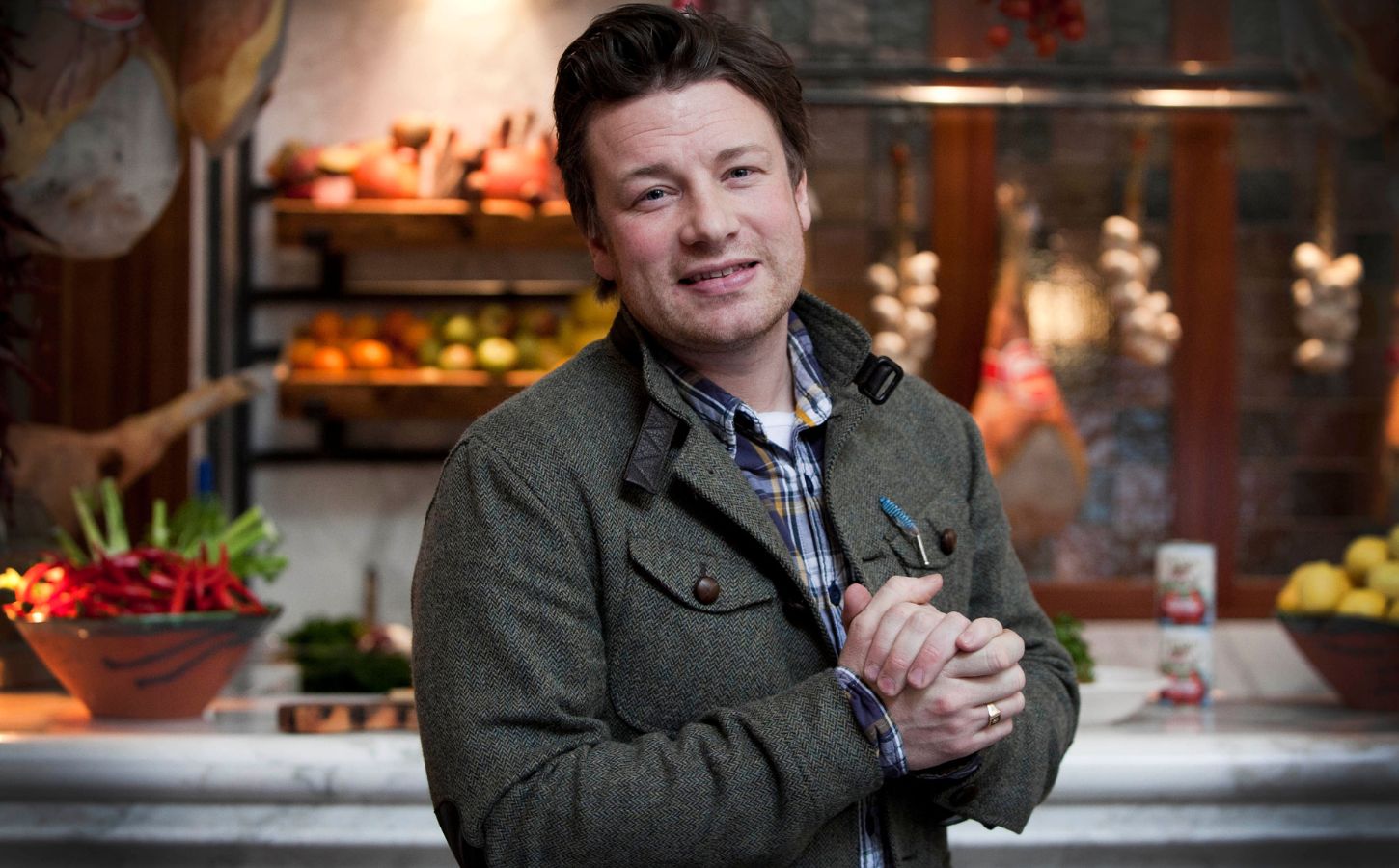 Photo shows Jamie Oliver standing in front of a kitchen stocked with fresh fruit and vegetables