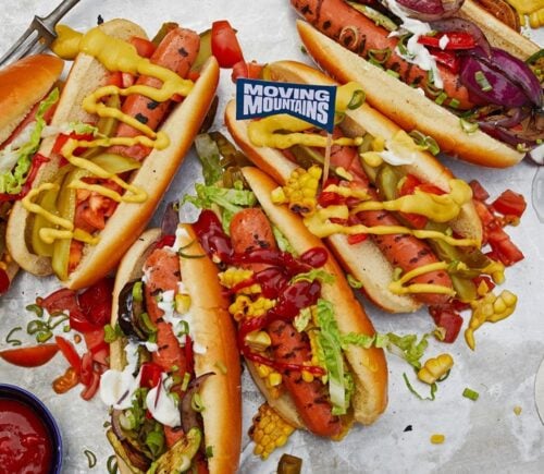 Photo shows a selection of vegan Moving Mountains hot dogs topped with pickles and sauce
