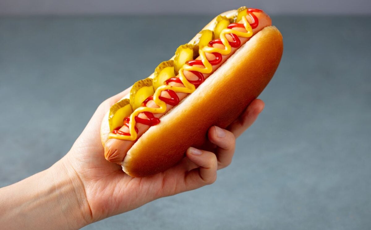 Photo shows someone's hand as they hold up a hotdog with sauces and pickles