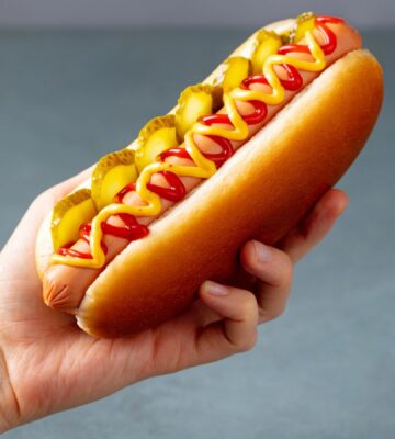 Photo shows someone's hand as they hold up a hotdog with sauces and pickles