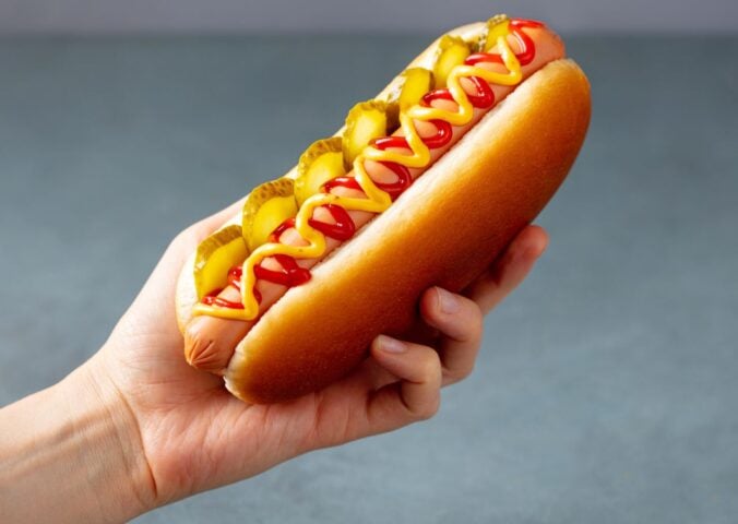 Photo shows someone's hand as they hold up a hotdog with sauces and pickles