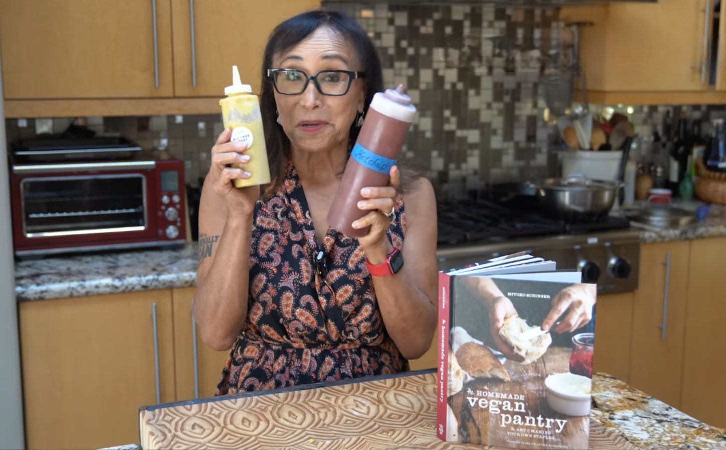 Miyoko Schinner stands in her kitchen holding two squeeze bottles of her homemade vegan ketchup and mustard