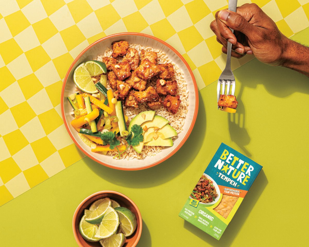Photo shows a bowl of cooked Better Nature tempeh alongside vegetables and product packaging while someone's hand holds a fork off to the side