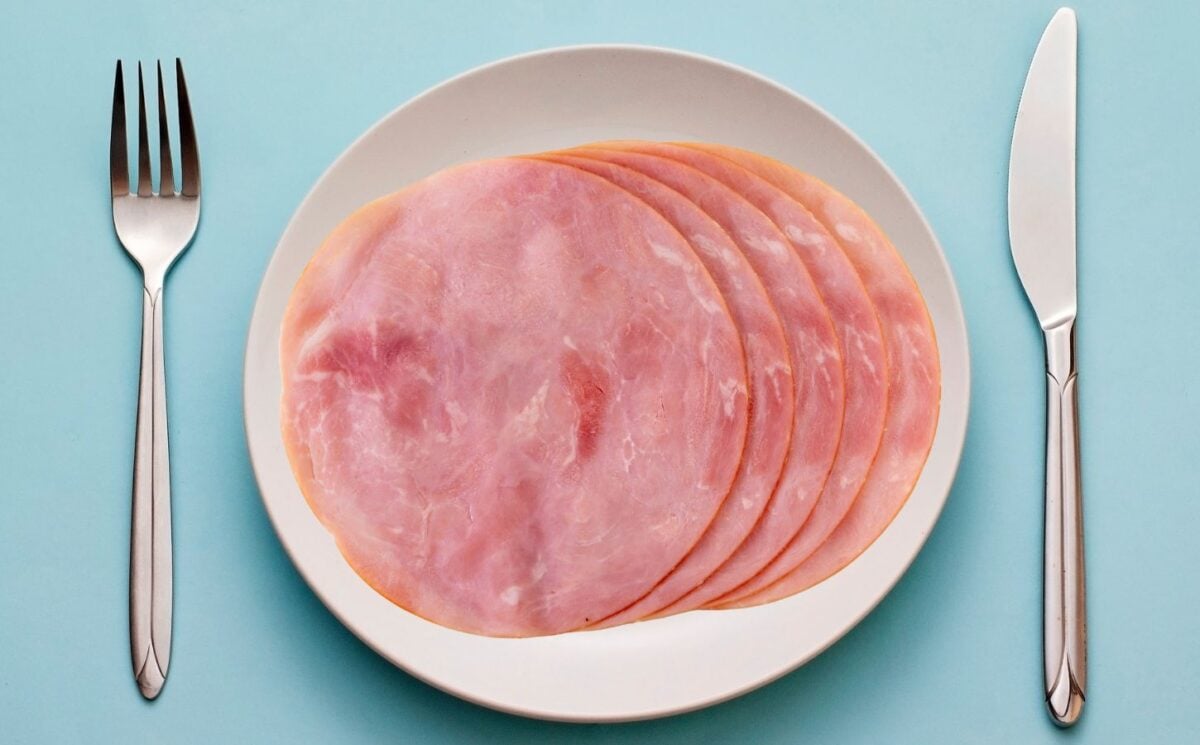 Photo shows a few slices of processed ham on a white plate against a blue background, flanked by cutlery