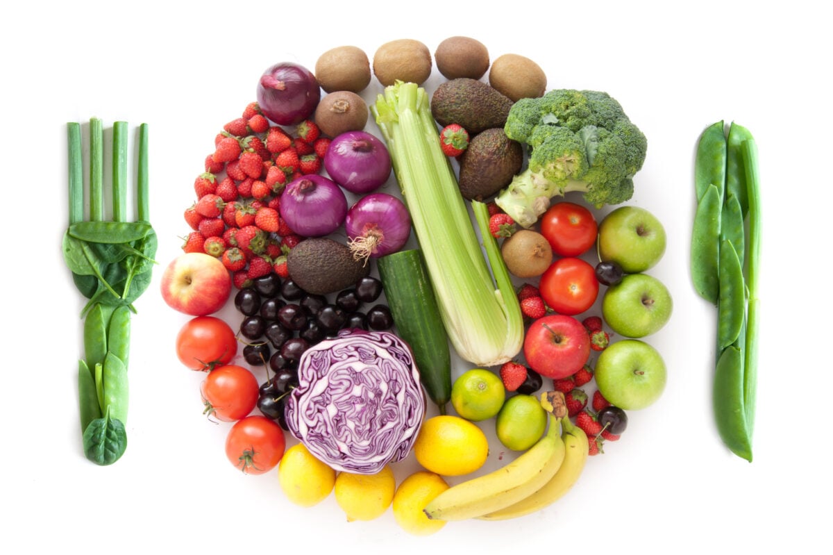 Photo shows a circle of fresh fruit and vegetables in the shape of a plate against a white background, next to a knife and fork made up of fresh greens and leaves