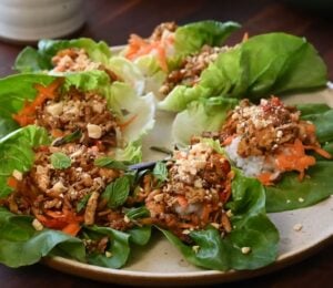 A plate of fresh lettuce wraps filled with a block of tofu, sushi rice, shredded carrots, herbs, and crushed peanuts