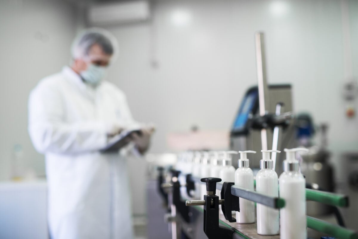 Photo shows someone in a white coat and mask, with a clipboard, inspecting bottles on a production line