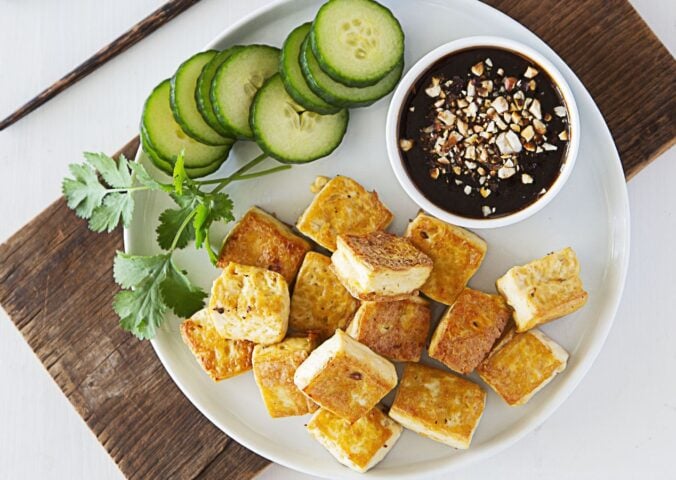 a plate of crispy tofu dipped in sweet and sour sauce with a side of cucumber