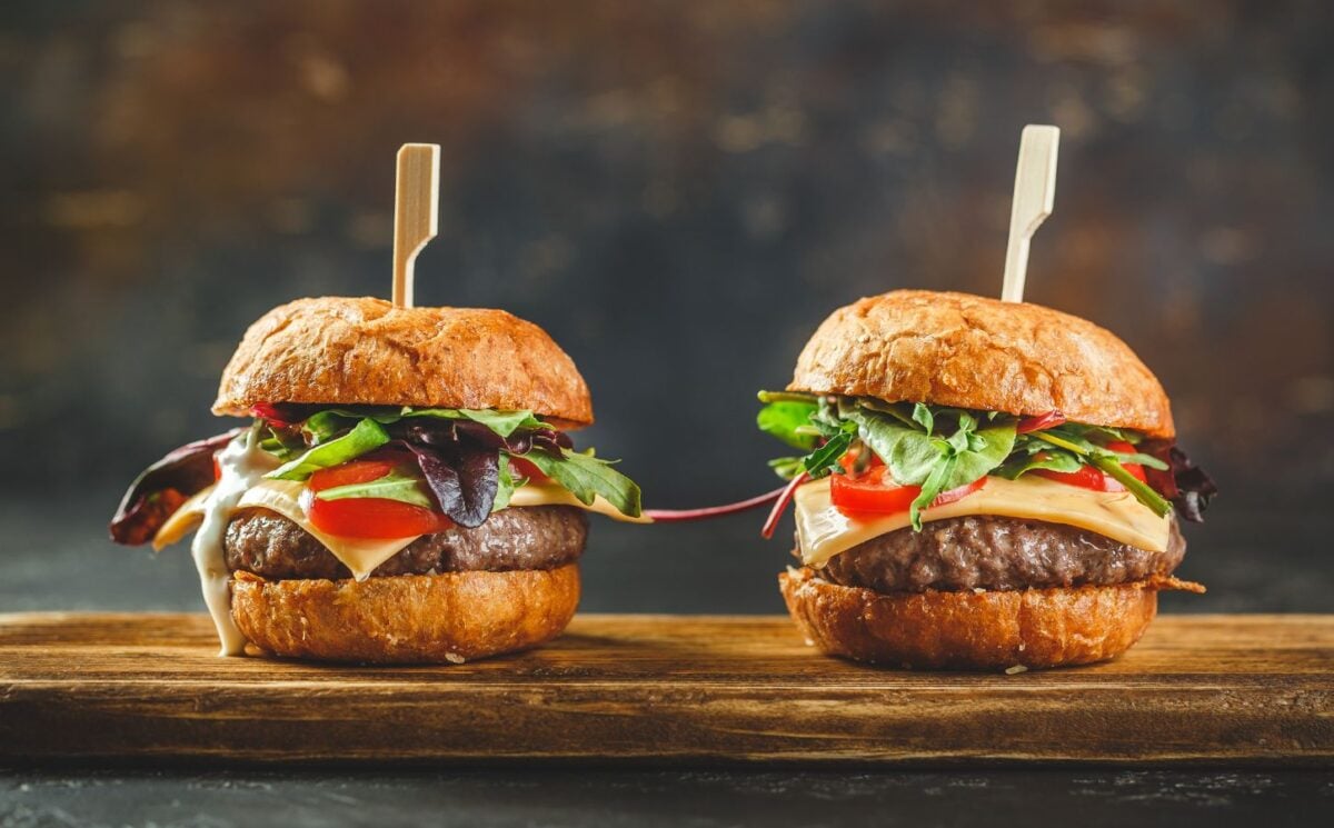 Photo shows two tall cheeseburgers side by side on a wooden board