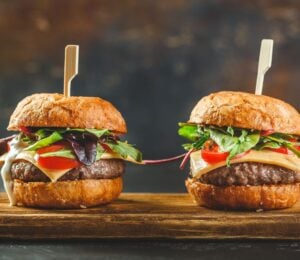 Photo shows two tall cheeseburgers side by side on a wooden board