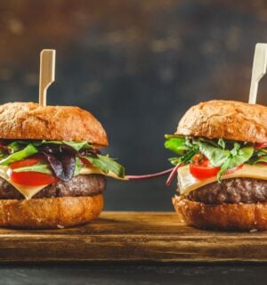 Photo shows two tall cheeseburgers side by side on a wooden board