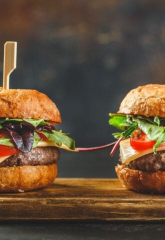 Photo shows two tall cheeseburgers side by side on a wooden board