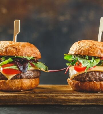 Photo shows two tall cheeseburgers side by side on a wooden board