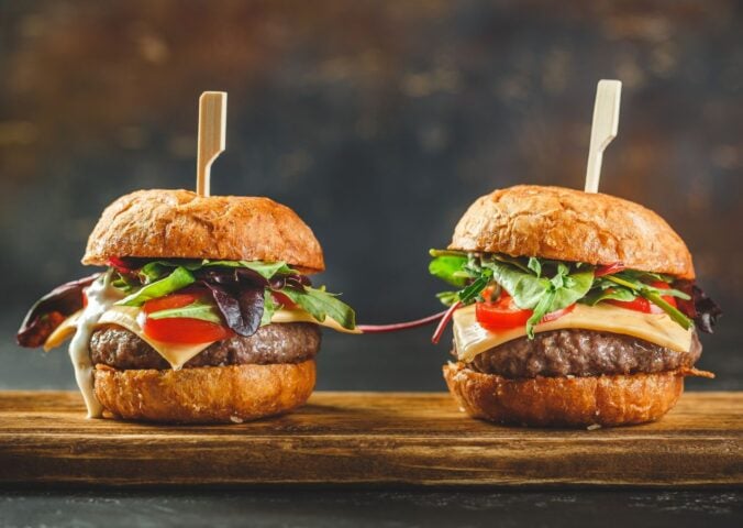 Photo shows two tall cheeseburgers side by side on a wooden board